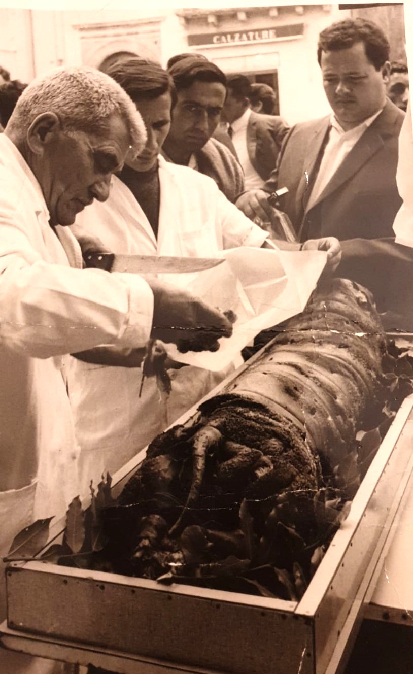 Fotografia storica del gesto artigianale durante la lavorazione della porchetta, contesto pubblico abruzzese, seconda meta del Novecento.